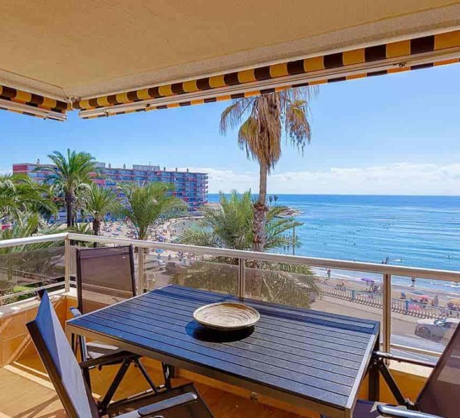 Photo d’un balcon donnant sur la mer à Torrevieja. Le balcon a un plancher en bois et un auvent rayé. Il y a une table et deux chaises sur le balcon. La vue depuis le balcon est celle de palmiers, d’une plage et de la mer. Le ciel est bleu et la mer est d’une couleur bleu clair. Il y a des bâtiments en arrière-plan.